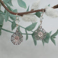 Silver earrings with intricate designs hanging from a branch with leaves and flowers.