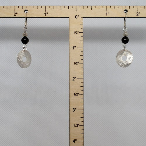 Pair of earrings with black beads and silver charms hanging from a ruler for scale on a gray background
