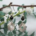 Pair of silver and black beaded earrings hanging on a branch with white flowers.