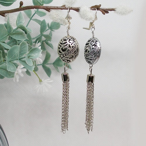 Silver earrings with intricate designs and tassels on a white background with green leaves.