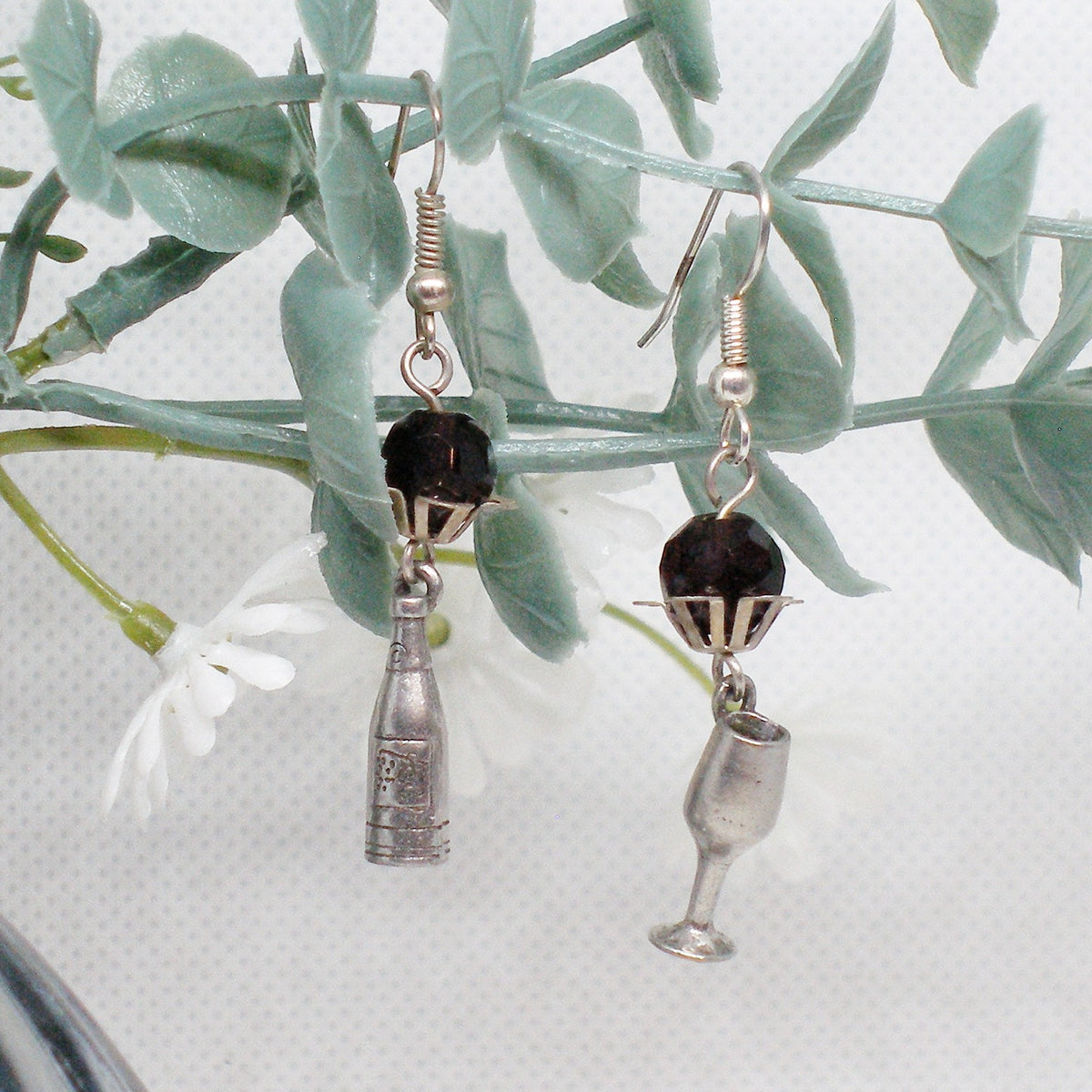 Pair of earrings with bottle and glass charms on a leafy branch.