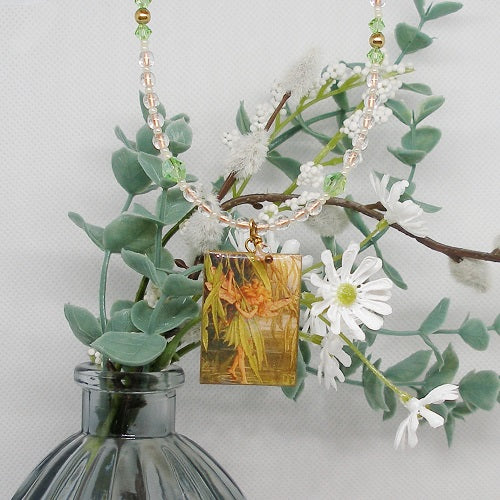 Necklace with a rectangular pendant featuring a fairy  and flowers, hanging over a glass bottle with greenery and white flowers.