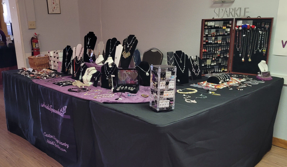 Table with jewelry display at a vendor event in a room with wooden floors.
