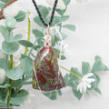 Necklace with a pendant featuring red and green colors, hanging against a natural background with green leaves and white flowers.