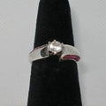 Silver ring with a white gem and ruby stones on a black stand against a gray background