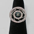 Silver ring with a central pearl and diamond-like stones on a black stand against a gray background