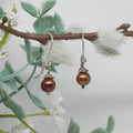 Pair of brown pearl earrings on a branch with green leaves and white flowers.