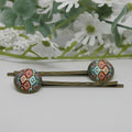Two hair pins with patterned ends on a white surface with flowers in the background