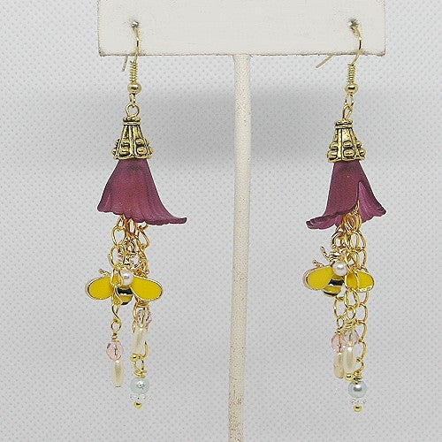 Pair of earrings with purple flower-like elements and gold bee accents on a white stand.