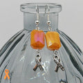Orange beaded earrings with silver ribbon charms on a glass bottle.
