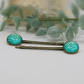 Two teal hair pins with gold accents on a white surface with green leaves in the background