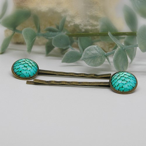 Two teal hair pins with gold accents on a white surface with green leaves in the background