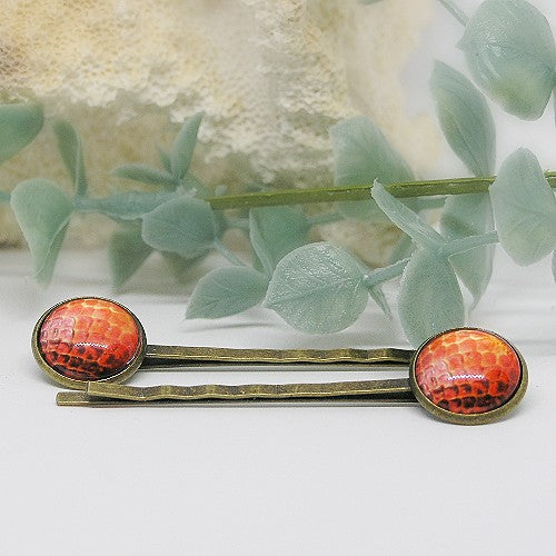 Two hair pins with orange and gold design on a white surface with green leaves in the background