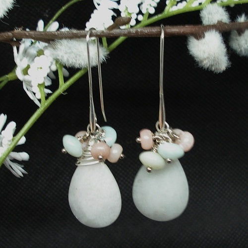 Pair of earrings with white teardrop stones and pink beads hanging on a branch with white flowers.