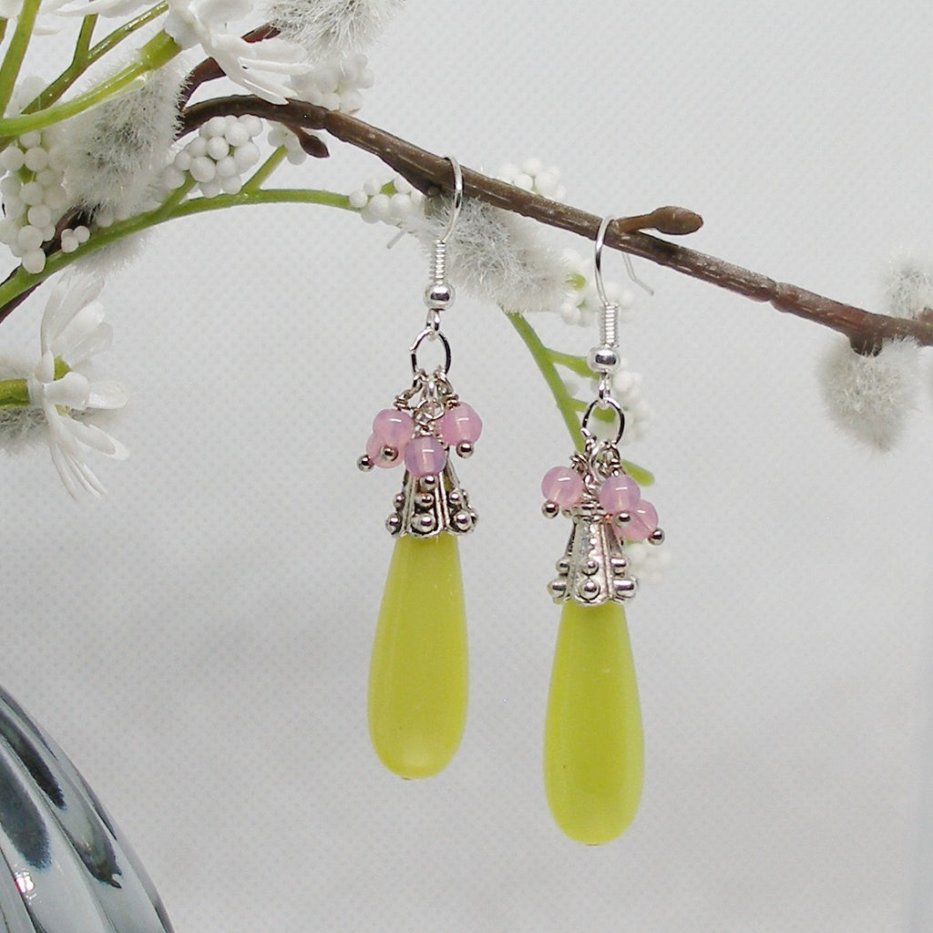 Green teardrop earrings with pink beads hanging from a branch with white flowers.