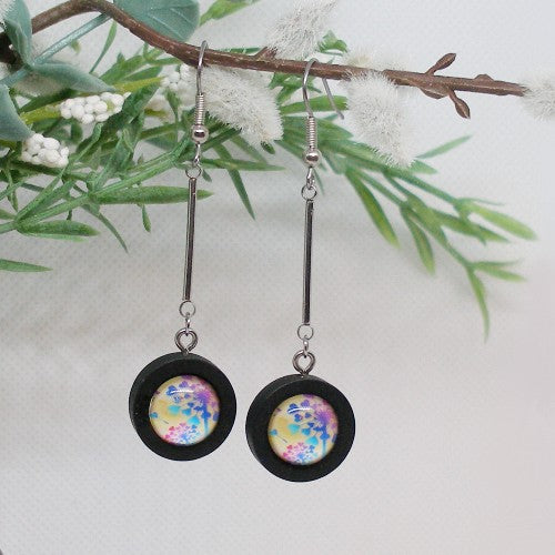 Pair of earrings with colorful circular designs hanging from a branch with green leaves and white flowers.
