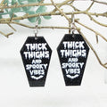 Black coffin-shaped earrings with white text and designs on a branch background