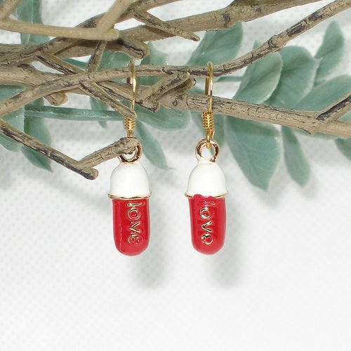 Red and gold capsule shaped earrings hanging on a branch with green leaves.