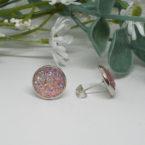 Round pink glittery earrings on a white surface with green leaves and white flowers in the background