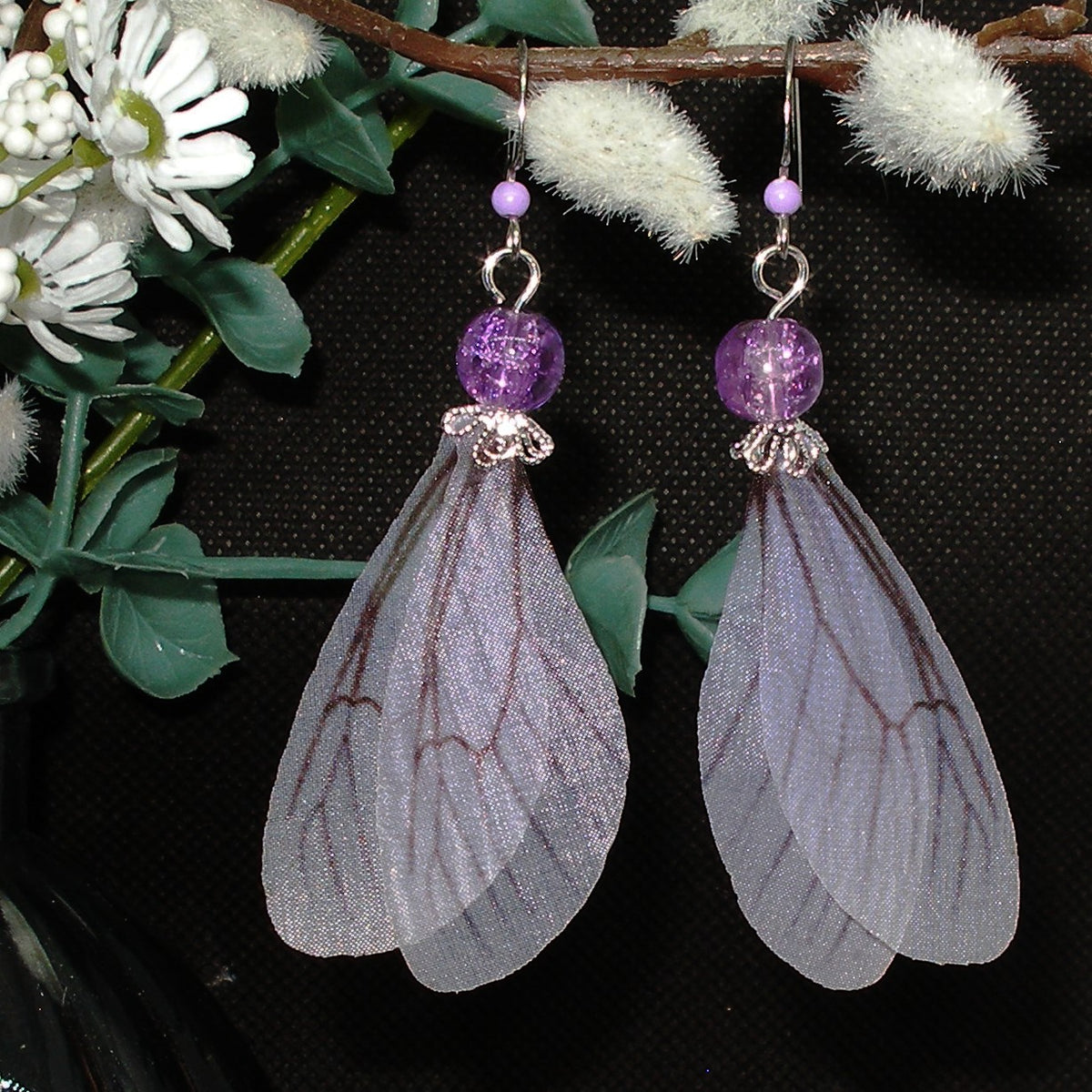 Pair of earrings with purple beads and sheer wing-like designs against a dark background.